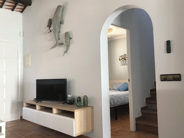 Ein Wohnzimmer mit einem Fernseher auf einem Holzboden, einer Wandskulptur und einer Tür zu einem Schlafzimmer mit Bett und Treppe.
