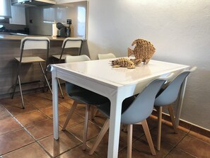 Ein Essbereich mit einem weißen Tisch und blauen Stühlen, im Hintergrund eine Küche und eine Tischskulptur aus gewebtem Korbmaterial.