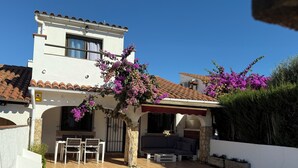 Ein zweistöckiges Haus mit einer gefliesten Terrasse, Sitzgelegenheiten und blühenden Pflanzen.