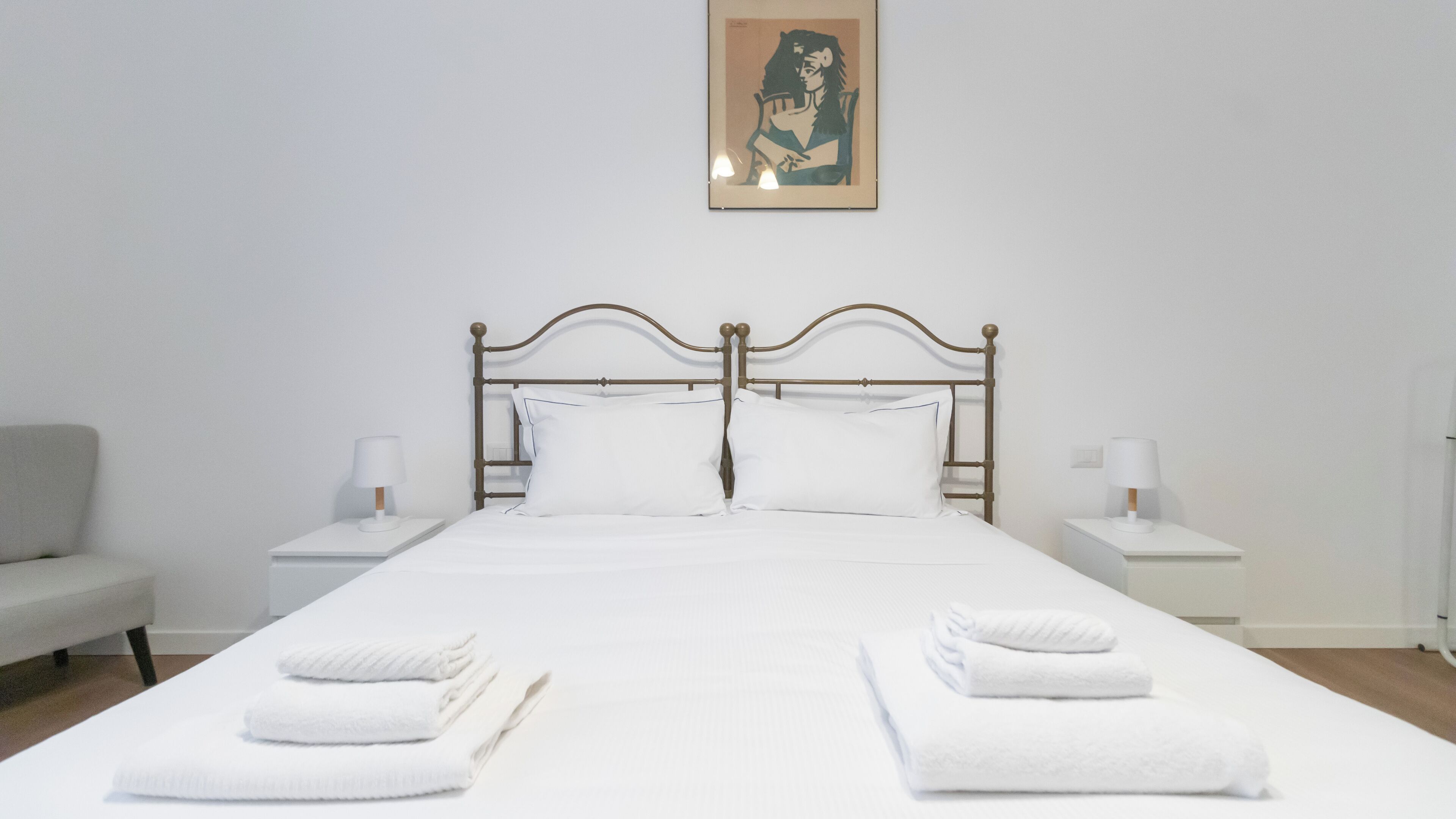 A neatly made bed with white linens and two pillows, flanked by white nightstands with lamps, in a minimalist room with a plain wall and a framed artwork above the bed.