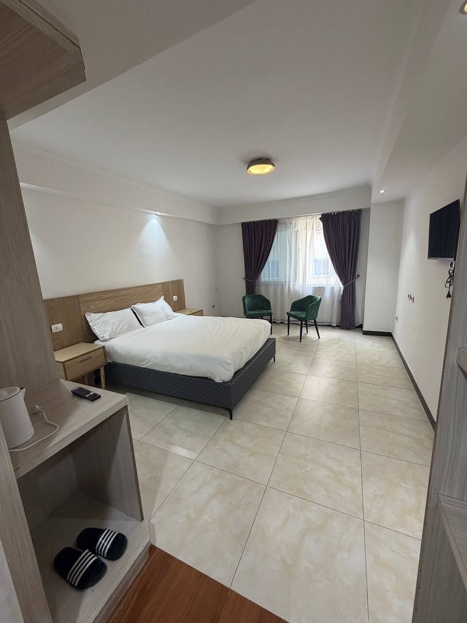 Hotelkamer met een bed, twee stoelen, een kleine tafel, een televisie en een plank met slippers.