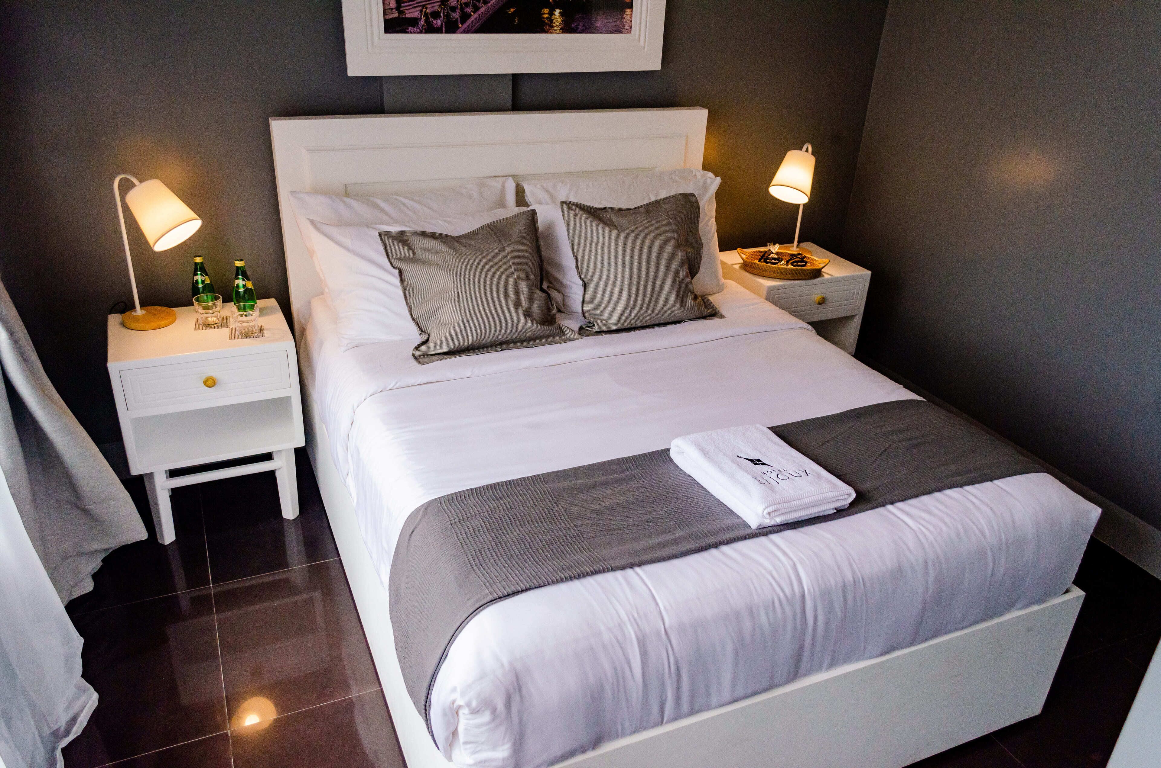 Una cama bien hecha con almohadas, una mesita de noche con una lámpara y una pequeña bandeja con botellas y vasos.