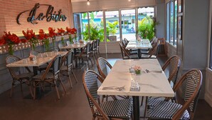 Ein Restaurant-Interieur mit Backsteinwänden, großen Fenstern und Korbstühlen. Die Tische sind mit Gläsern und Besteck gedeckt.
