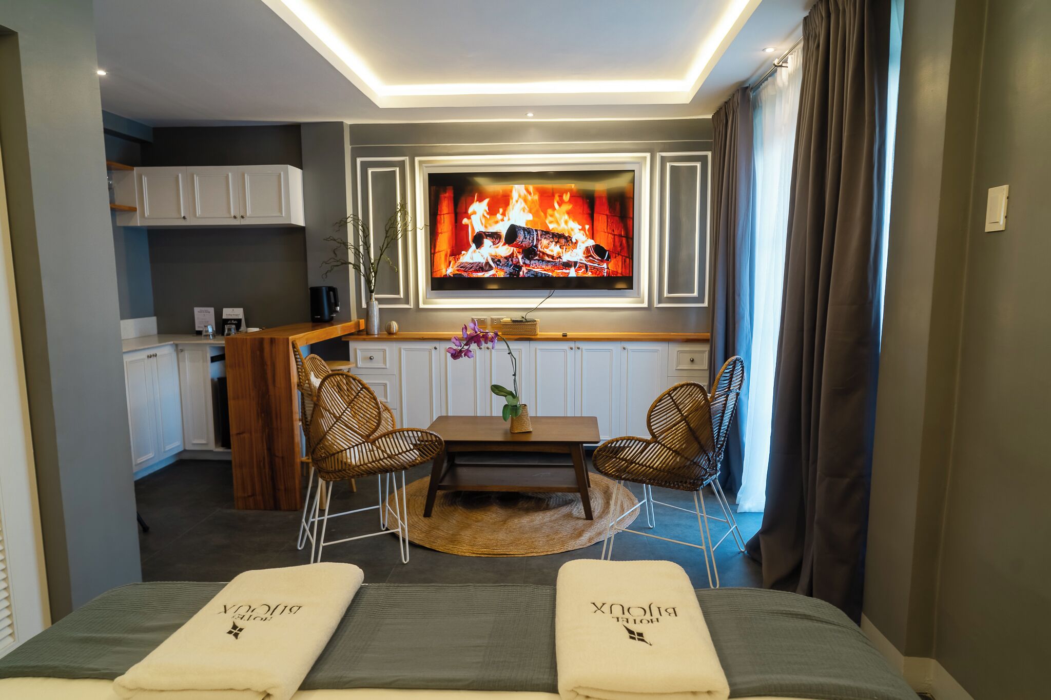 A hotel room with a bed, two chairs, a small table, a TV with a fireplace display, a kitchen area, and a bathroom.
