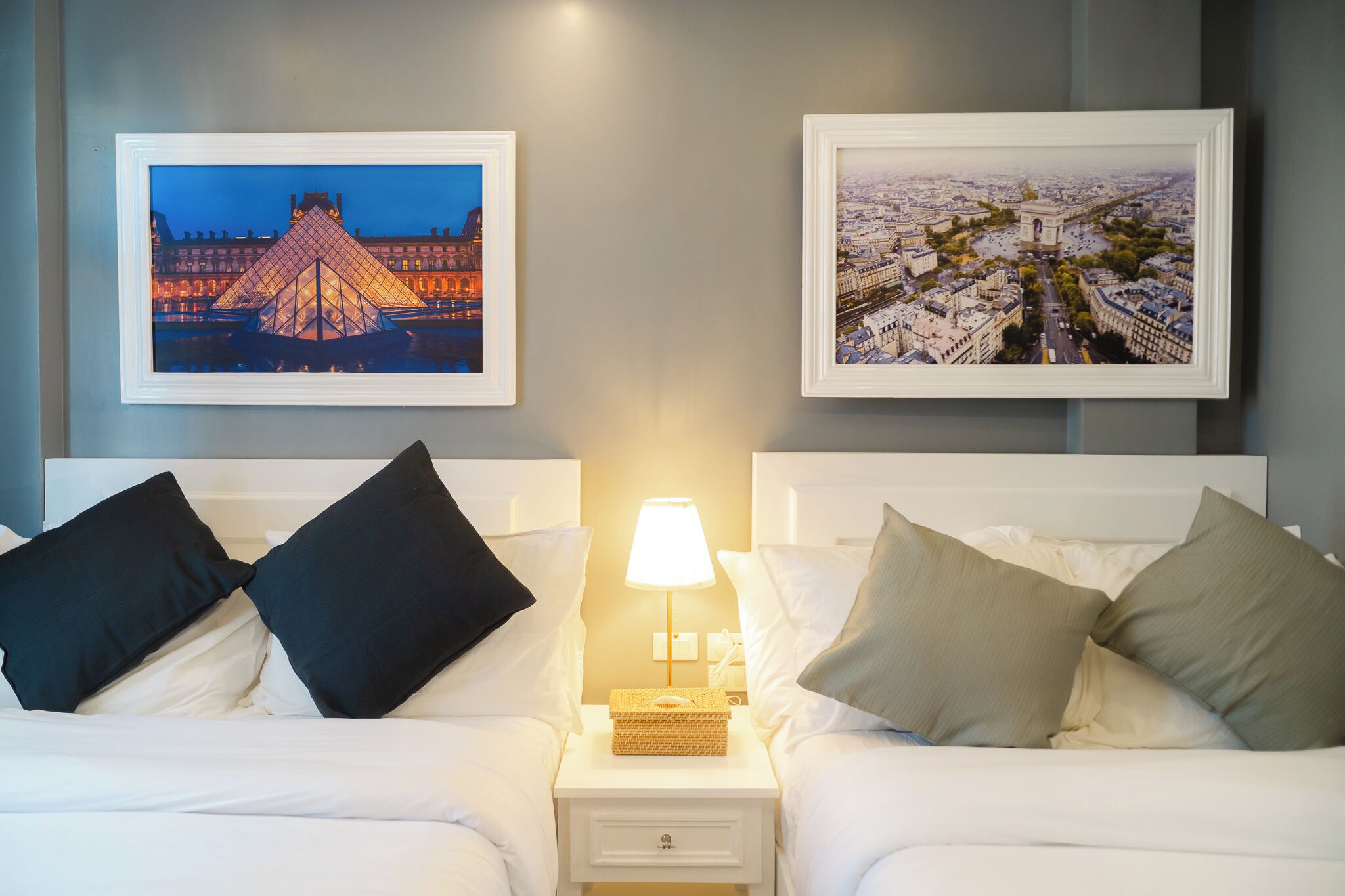 A hotel room with two beds, a nightstand with a lamp, and two framed pictures on the wall.