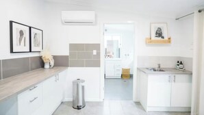 A modern kitchen with white cabinets, a grey countertop, and a trash bin. There are framed pictures on the wall and a basket on the countertop. - Pink Muse, 3 Mins to Ettalong Beach (Ettalong Beach)