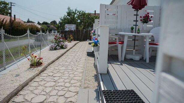 A covered outdoor seating area with a table and chairs, a small garden with flowers, and a white fence. - EHM: Modern Apartment Near Spa & Nature-A1 (Mesteri)