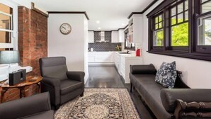 Ein modernes Wohnzimmer mit einer schwarzen Ledersofa, einem gemusterten Teppich und einer Wand mit Ziegeln. Im Hintergrund ist eine Küche mit weißen Schränken und einer schwarzen Spritzschutzwand zu sehen.