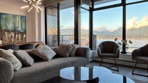 A modern living room with a large sofa, a glass coffee table, and a view of a cityscape through large windows. - Infinite blue (Vancouver)