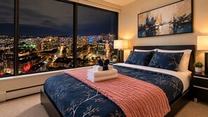 Un dormitorio con un ventanal grande, una cama con colcha azul y blanca, un plaid rosa y vistas nocturnas de la ciudad.