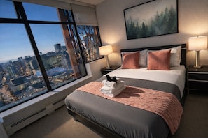 A hotel room with a large window, a bed with a pink blanket, two bedside lamps, and a cityscape view.