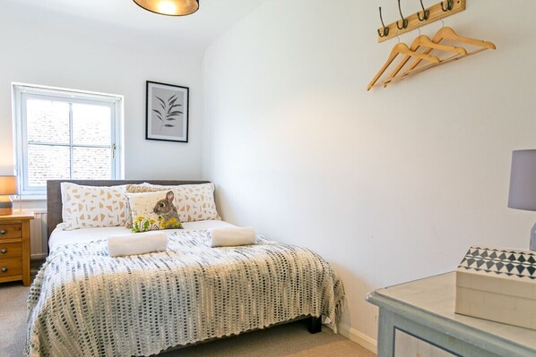 Een slaapkamer met een bed, nachtkastje, lamp, raam en een kapstok aan de muur.