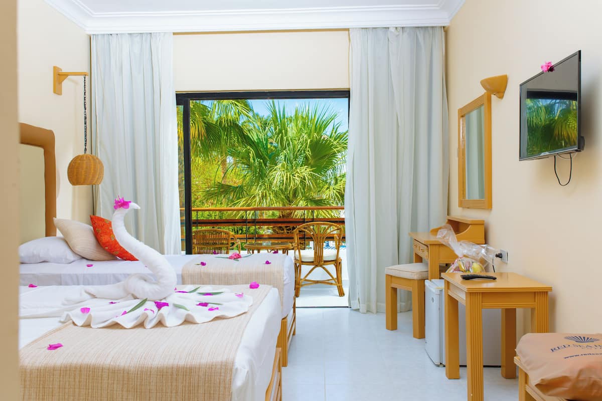 A hotel room with two beds, a balcony with a view of palm trees, a TV, and a desk with a chair.