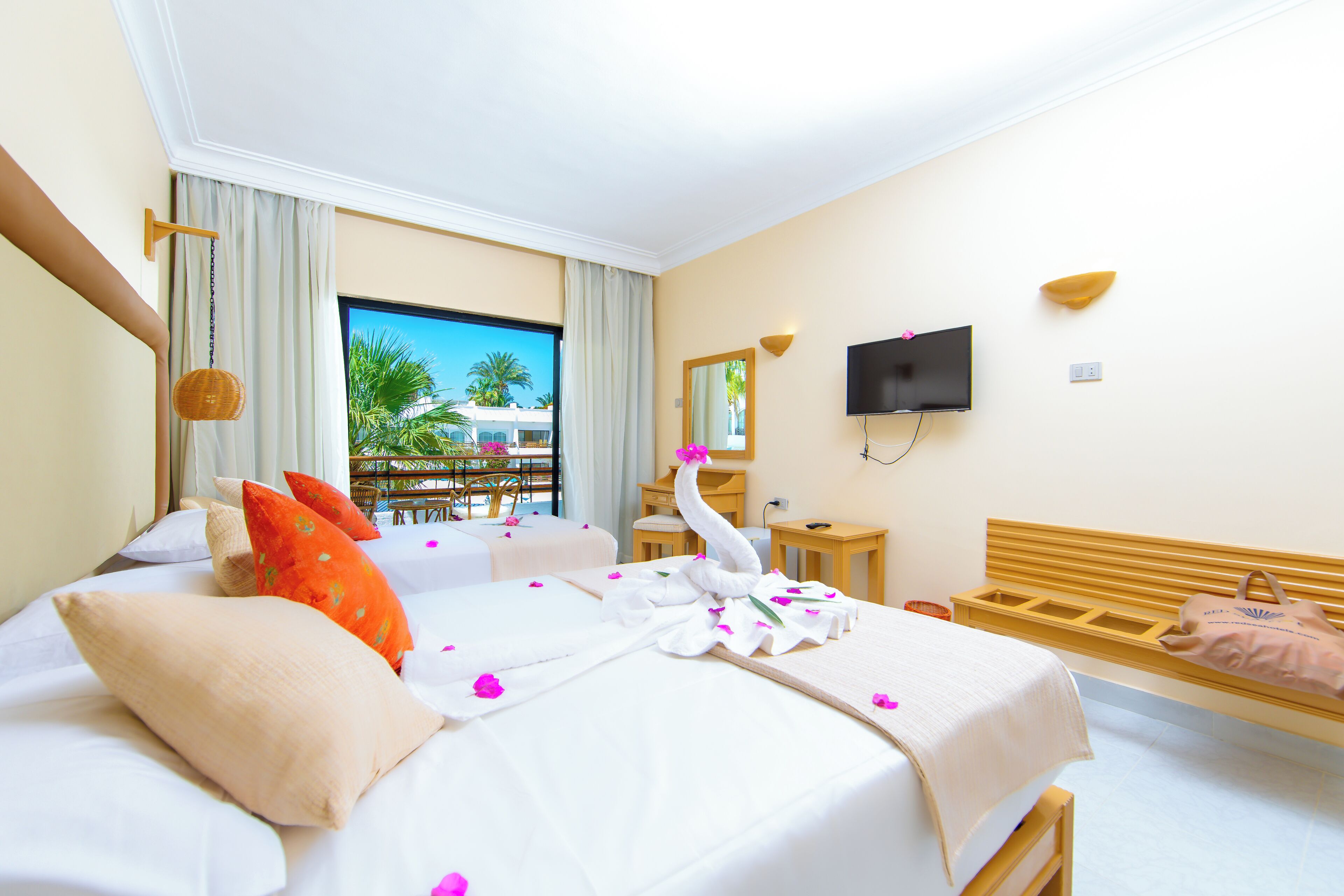 Una habitación de hotel con cama, televisión, cabecera de madera, ventana con vista a palmeras y balcón.