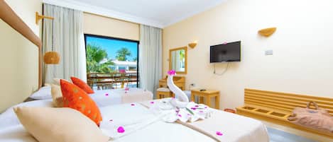 Una habitación de hotel con cama, televisión, cabecera de madera, ventana con vista a palmeras y balcón.