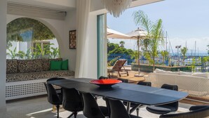 A modern dining area with a large table, black chairs, and a red bowl. There is a patterned sofa with green cushions and a view of the outdoors through a large window. - Dar Elyssa (Sidi Bou Said)