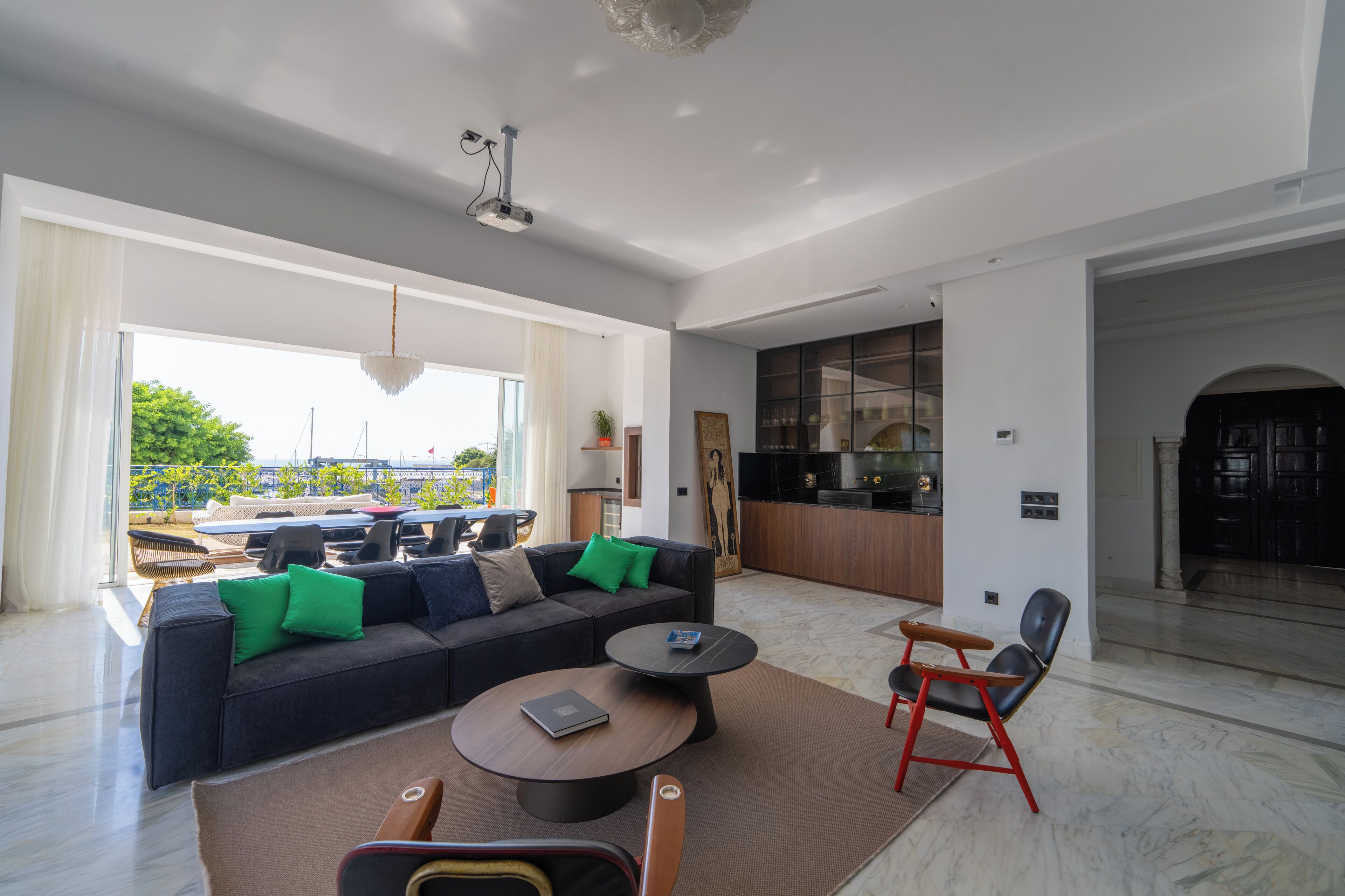 A modern living room with a sofa, coffee table, and chairs, a kitchen area, and a view of the outdoors.