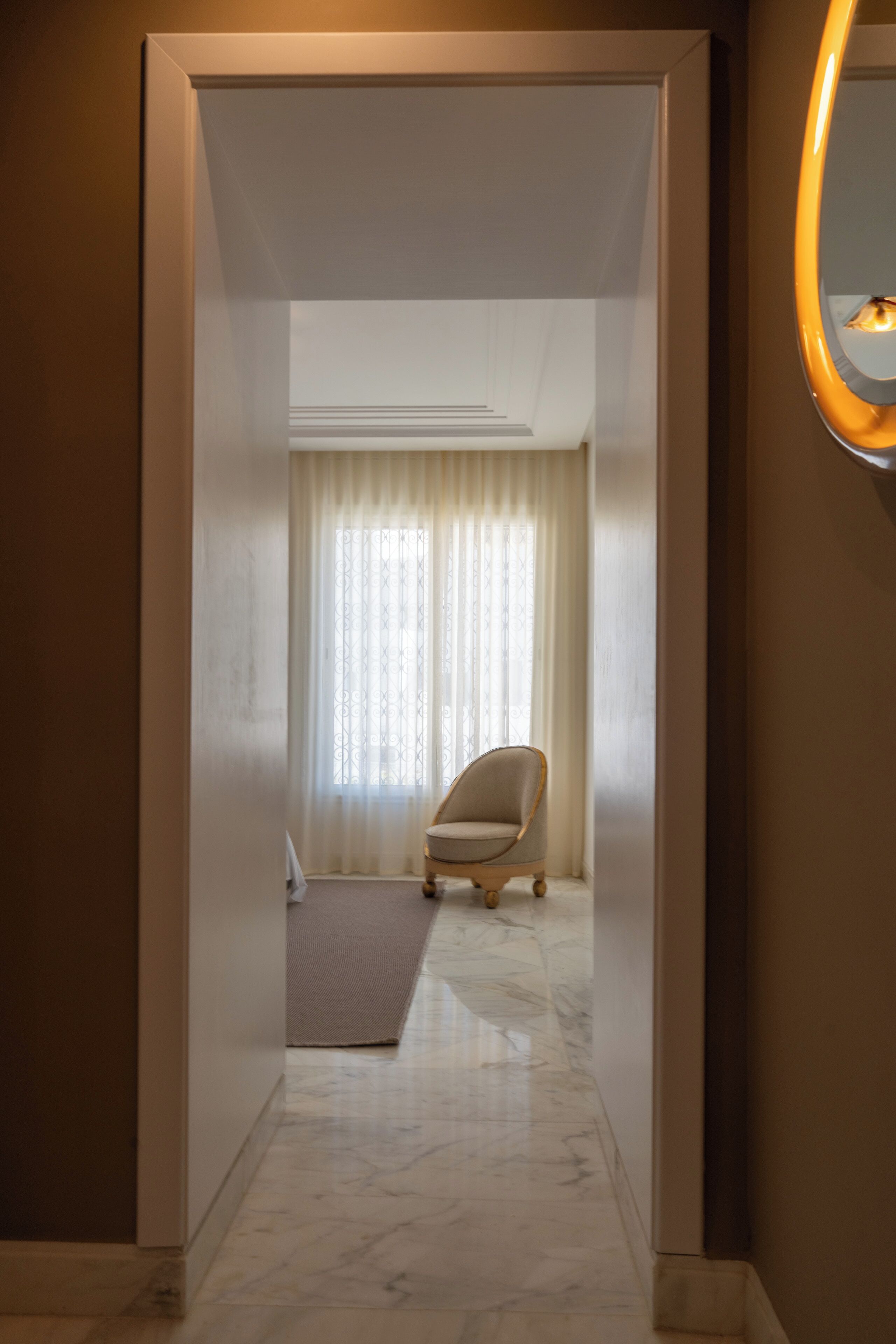 A hallway with a marble floor leading to a room with a chair and a window with curtains.