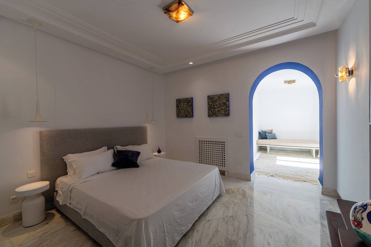 A bedroom with a large bed, a bedside table, a white wall, and a blue archway leading to a balcony.