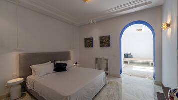 A bedroom with a large bed, a bedside table, a white wall, and a blue archway leading to a balcony.