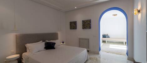 A bedroom with a large bed, a bedside table, a white wall, and a blue archway leading to a balcony.