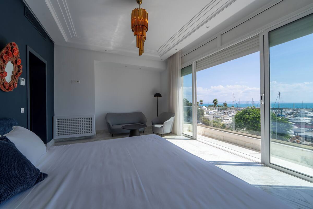 A hotel room with a large bed, a sofa, a small table, and a view of the marina.