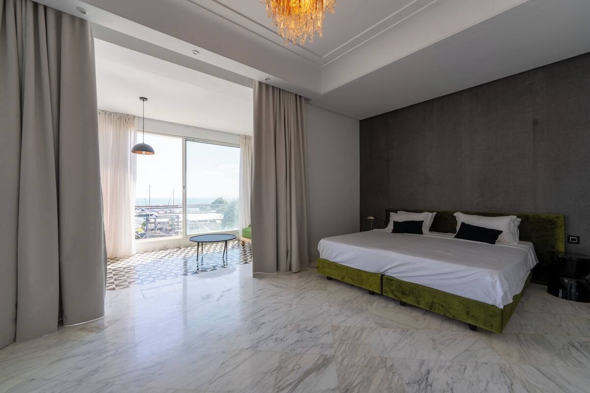 A spacious bedroom with a large bed, a balcony with a table, and a view of the city.
