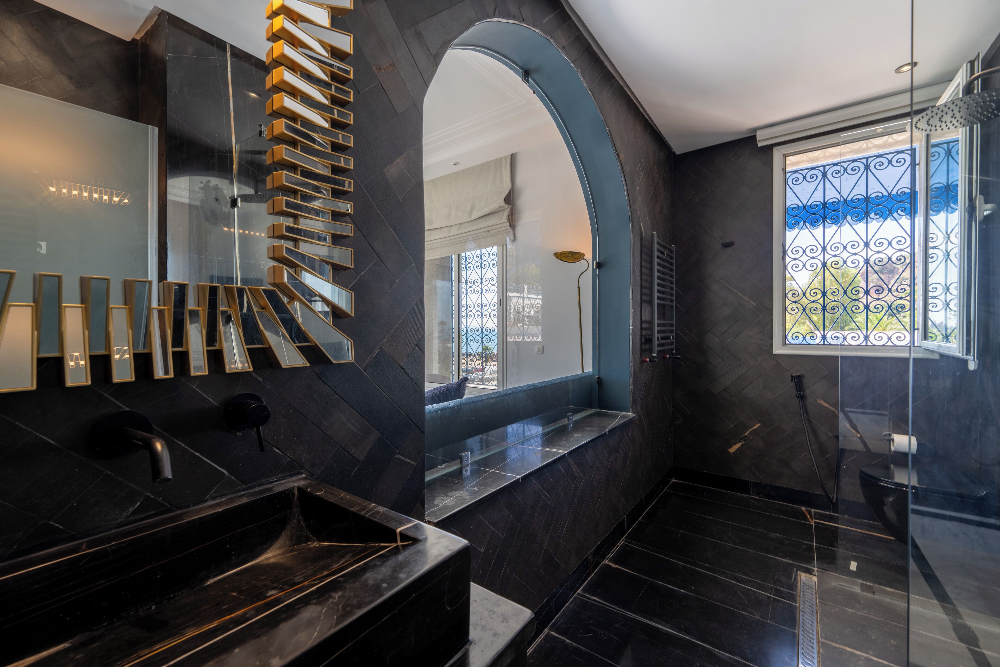 Casa de banho moderna com lavatório de mármore preto, cabine de duche em vidro e vista para o exterior através de uma janela com grades de ferro decorativas.