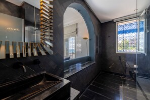 A modern bathroom with a black marble sink, a glass shower enclosure, and a view of the outdoors through a window with decorative iron bars. - Dar Elyssa (Sidi Bou Said)
