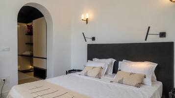 Un dormitorio moderno con una cama grande, ropa de cama blanca y un cabecero oscuro. Hay dos luces fijadas a la pared y una repisa con toallas dobladas.