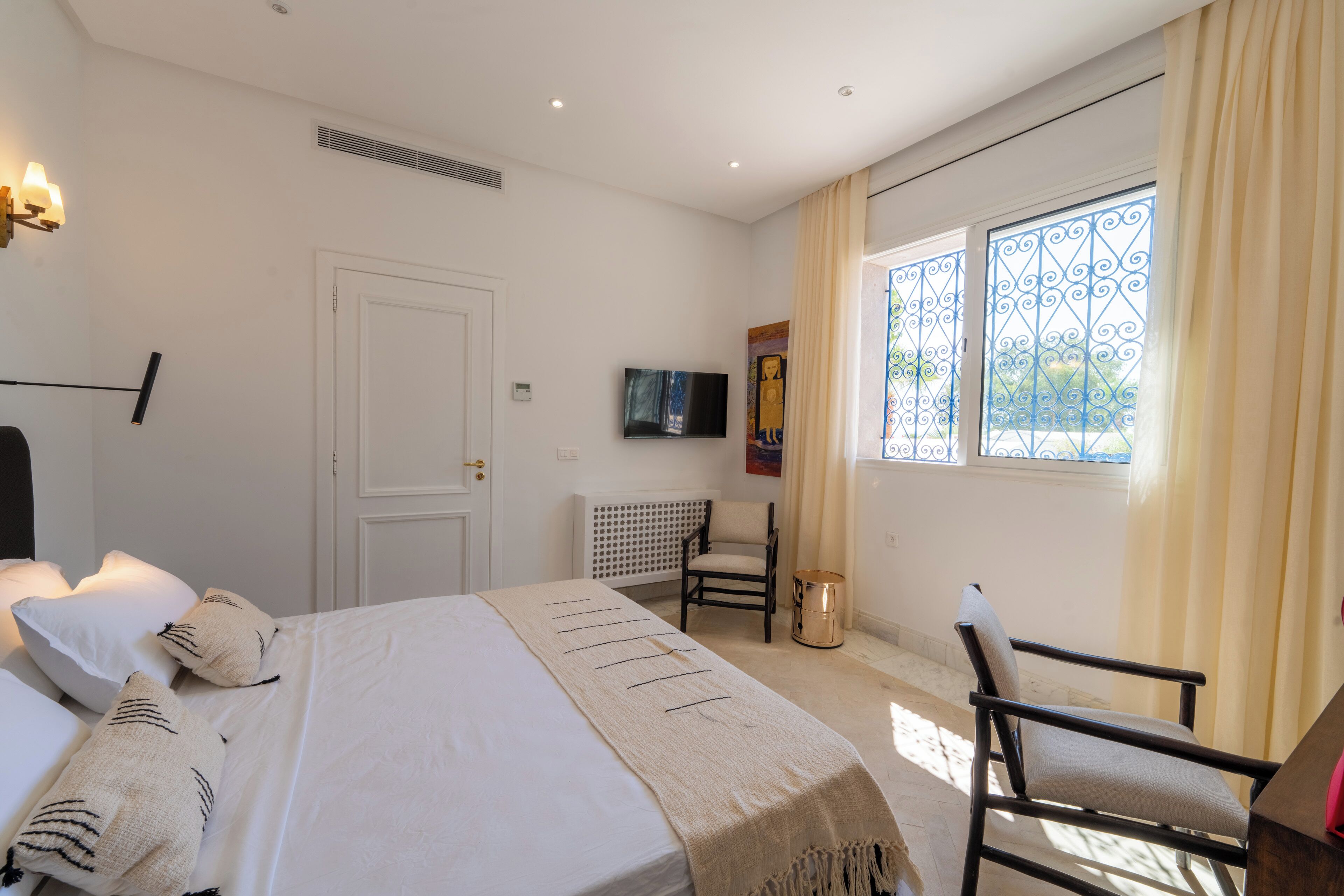 Un dormitorio con cama, una silla, un televisor y una ventana con cortinas.