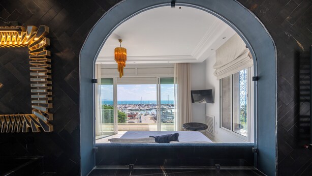 A bedroom with a large window, a bed, a TV, and a chandelier. - Dar Elyssa (Sidi Bou Said)