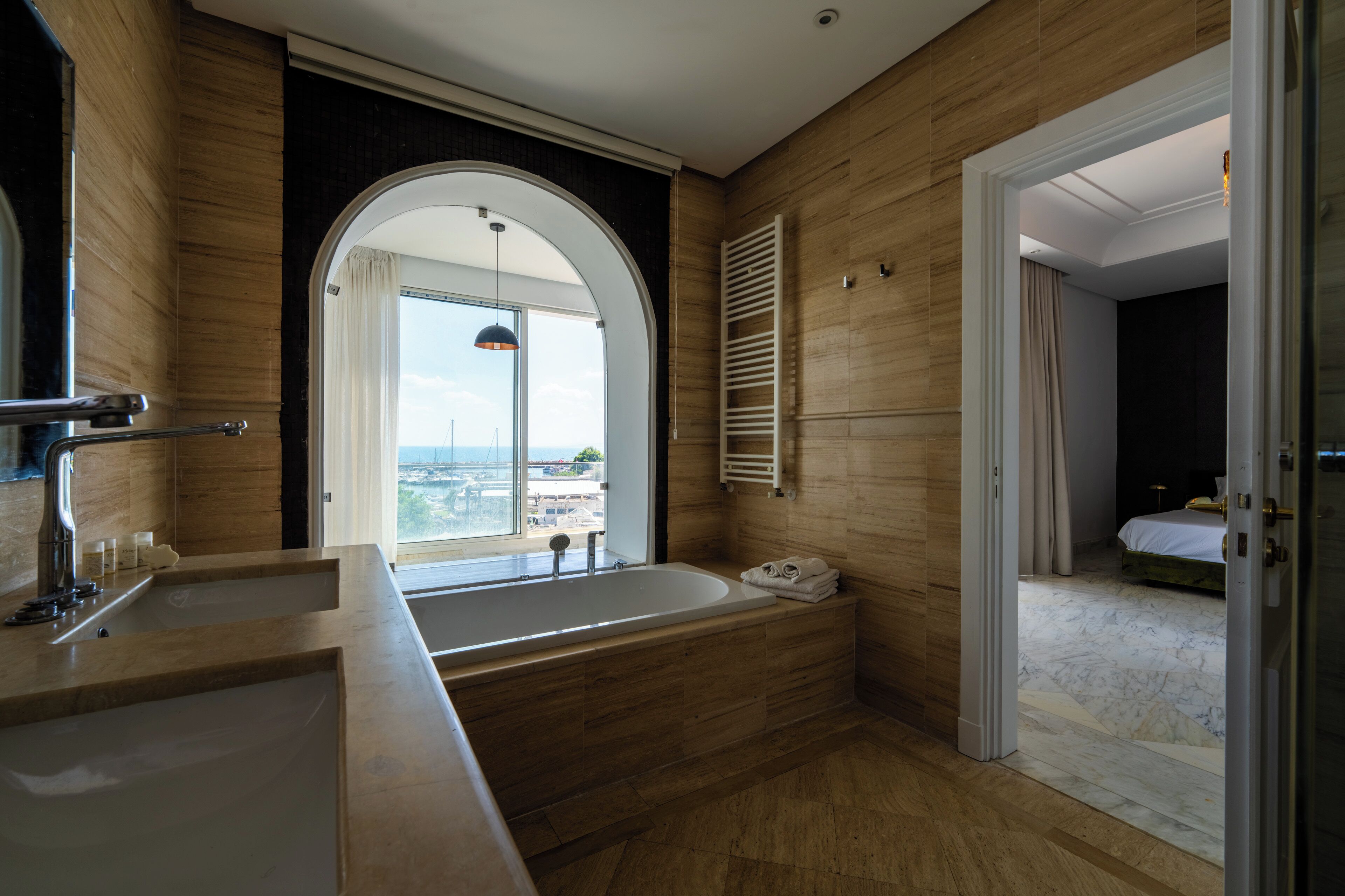 A modern bathroom with a large bathtub, a double sink, and a view of the city through an arched window.