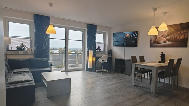 A modern living room with a sofa, coffee table, and dining area with chairs. There are large windows with curtains, a flat-screen TV, and a painting on the wall. - Guest apartment and service apartment Hermsdorf (Hohe Börde)