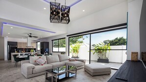 A modern living room with a sectional sofa, a glass coffee table, and a dining area with a kitchen in the background. - Modern 5 Bedrooms Pool Villa Curtis (Rawai)
