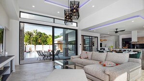 A modern living room with a large sofa, a glass dining table, and a view of a pool and outdoor area. - Modern 5 Bedrooms Pool Villa Curtis (Rawai)