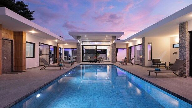 A modern indoor swimming pool with lounge chairs and a dining area in the background. - Modern 5 Bedrooms Pool Villa Curtis (Rawai)