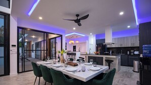 A modern kitchen with a central island, a dining area with green chairs, and a ceiling fan. - Modern 5 Bedrooms Pool Villa Curtis (Rawai)
