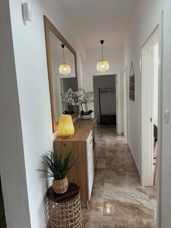 A hallway with a wooden console table, a mirror, hanging pendant lights, a potted plant, and a tiled floor.