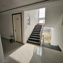 Uma escadaria com corrimão que leva a um andar superior, uma porta com placa de número e uma planta em vaso no chão.