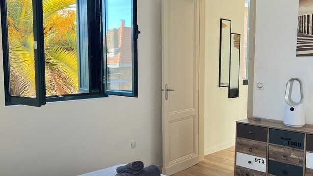 A bedroom with a bed, a window with an open view of palm trees, a wooden dresser, and a door leading to another room. - Apartment Arcachon Centre "Escale sur le Bassin" (Stopover on the Basin) (ARCACHON)