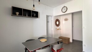 Ein moderner Essbereich mit einem grauen Tisch, roten Sitzhussen, einer Pendelleuchte und einem an der Wand befestigten Regal mit Dekorationselementen.