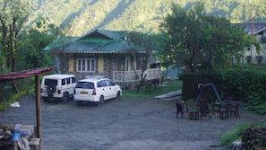A rustic building with a green roof, surrounded by lush greenery and parked vehicles.