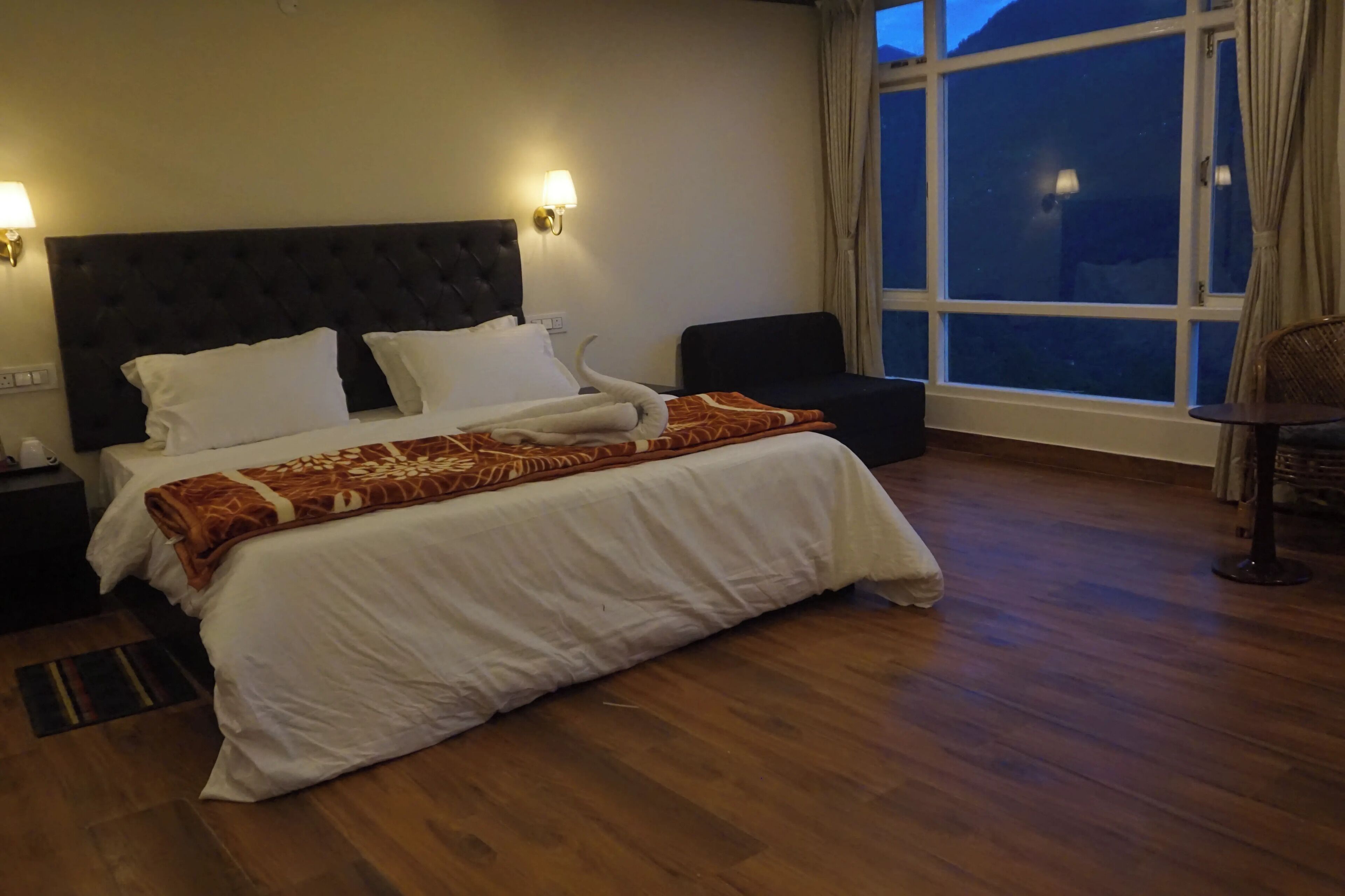Una habitación de hotel con una cama grande, dos lámparas de noche, una silla y un ventanal con cortinas.