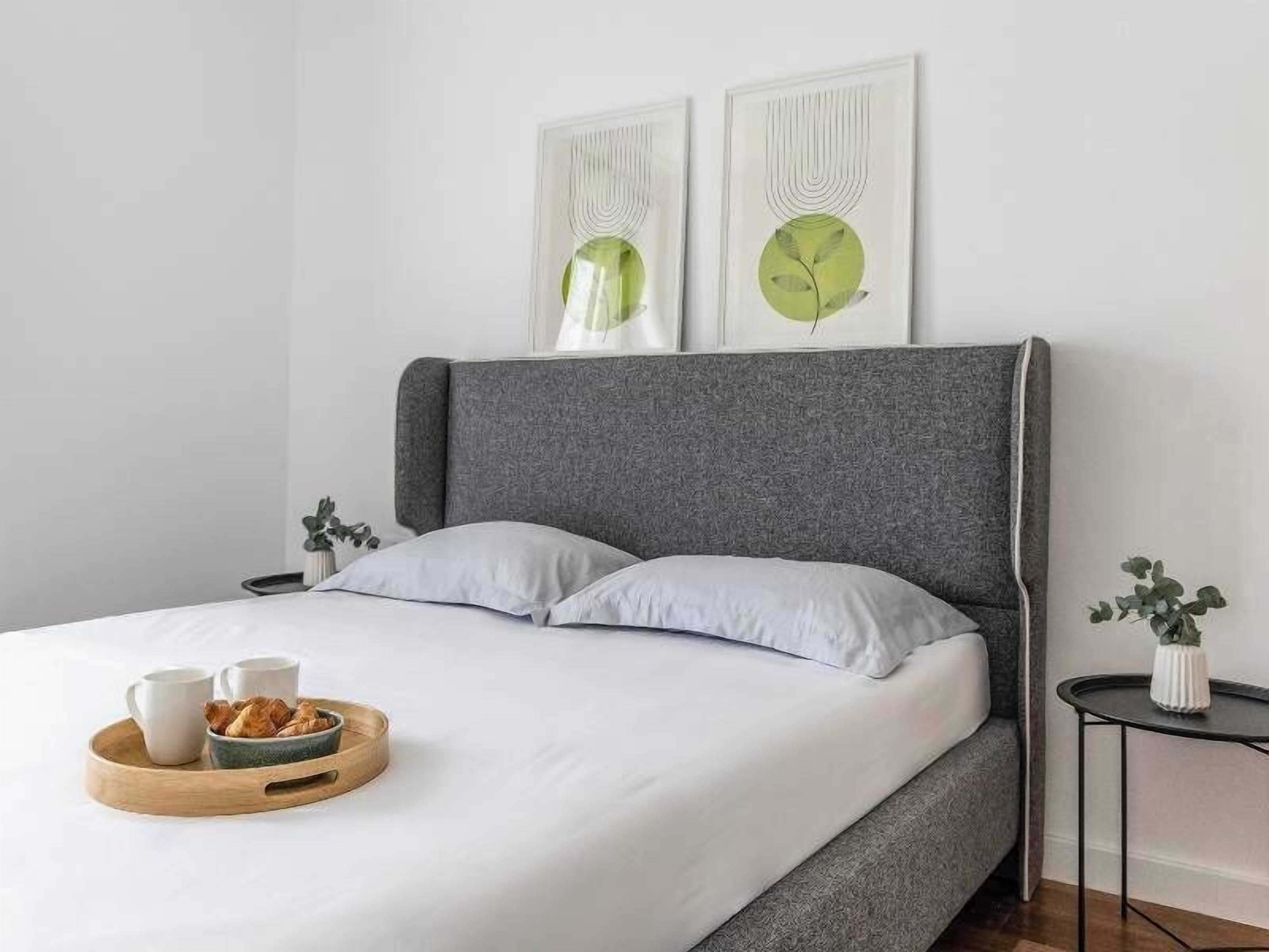 Een moderne slaapkamer met een grijs gestoffeerd hoofdbord, witte beddengoed en een houten dienblad met ontbijtartikelen.