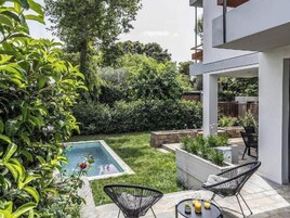 Un patio moderno al aire libre con piscina, rodeado de exuberante vegetación y un edificio blanco.
