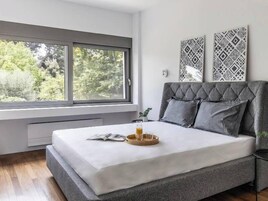 Un dormitorio moderno con un ventanal grande, una cama con ropa de cama gris y dos cuadros decorativos con diseños.