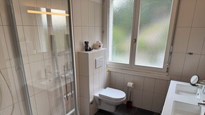 Et badeværelse med glasvæg omkring bruseniche, toilet, håndvask og et vindue med satin glas.