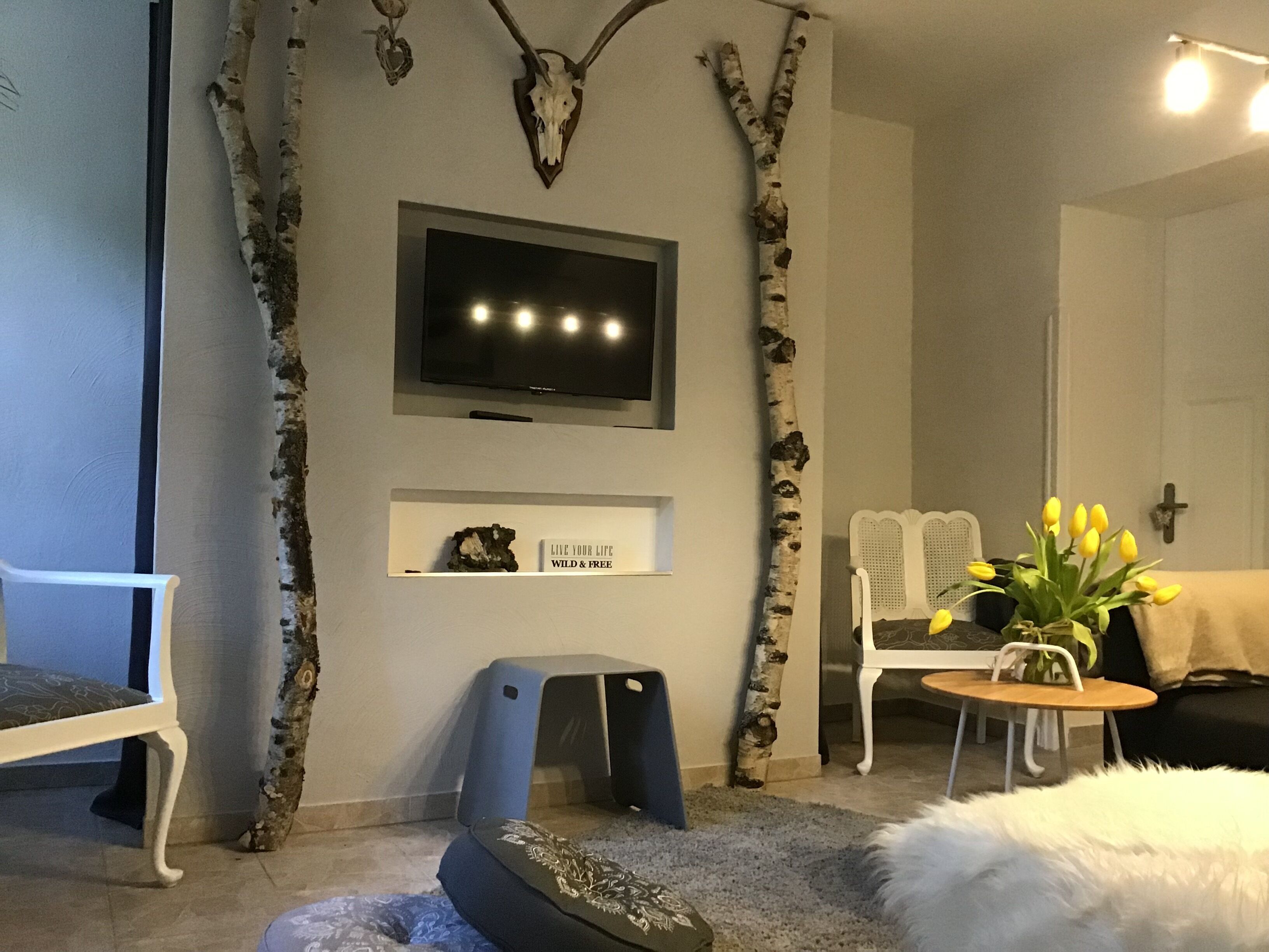 A living room with a mounted TV, a white sofa, a birch tree branch, a small table with a vase of flowers, and a cross on the wall.