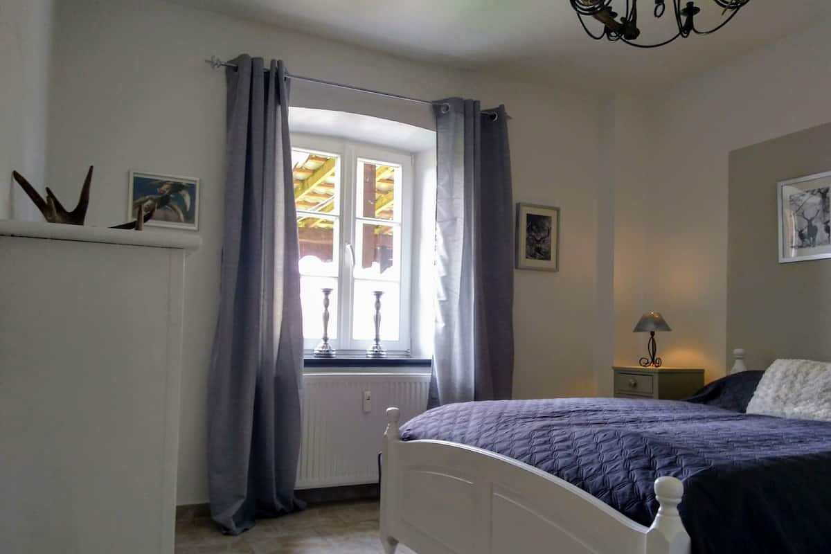 A bedroom with a bed, a window with curtains, a radiator, a nightstand with a lamp, and wall art.